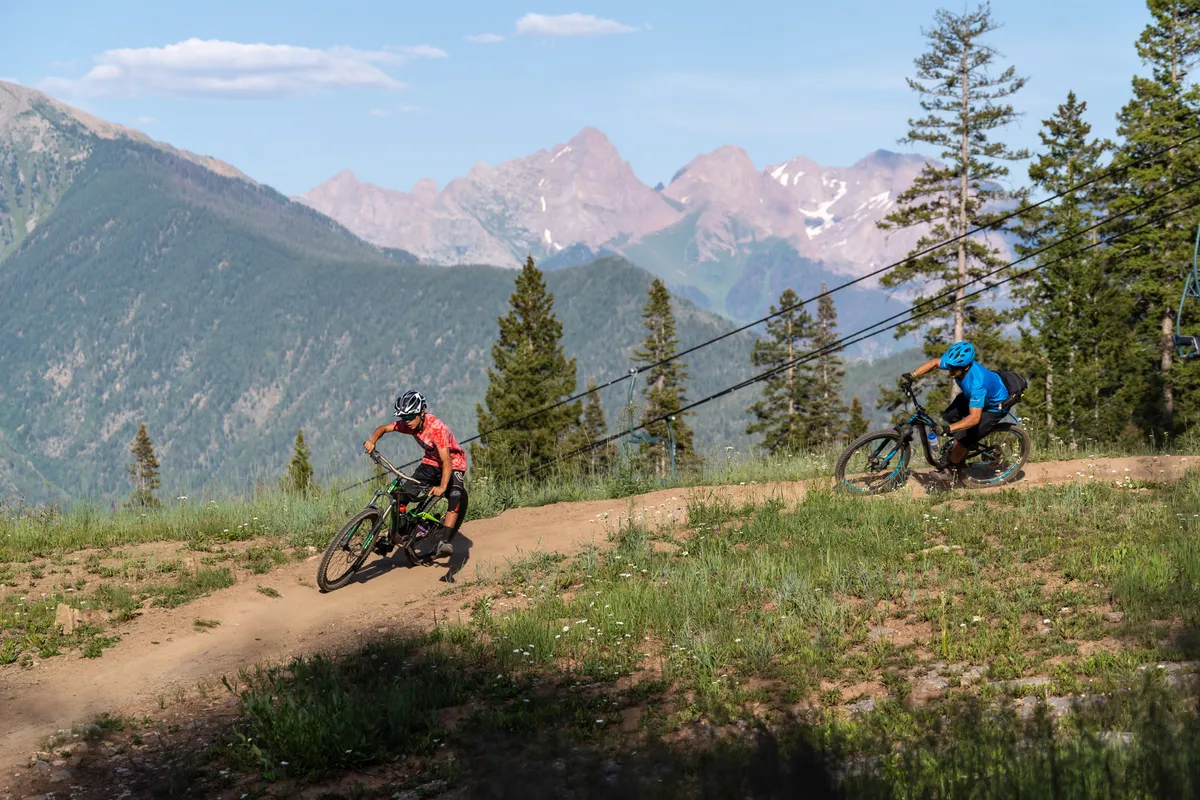 This image captures mountain bikers navigating a dirt trail surrounded by lush alpine trees and set against a dramatic mountain backdrop beneath a clear blue sky. The scene is energetic and vibrant, reflecting the active outdoor pursuits available in the area.