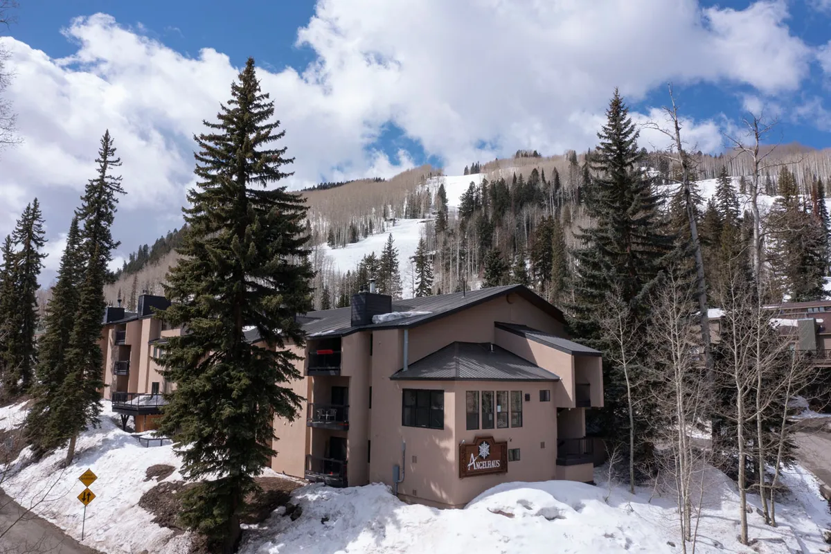 A tranquil exterior view featuring a snow-covered landscape, tall pine trees, and the architecture of nearby lodges set against a mountainous backdrop. The setting is peaceful, highlighting both the natural beauty and the convenience of lodging close to the slopes.