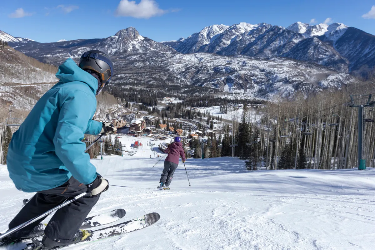 This image captures skiers in action on a snowy slope, with a sweeping mountain range and a ski resort visible in the valley below. The scene is lively with people actively skiing and snowboarding, while the expansive winter landscape highlights the proximity to the slopes and resort amenities.