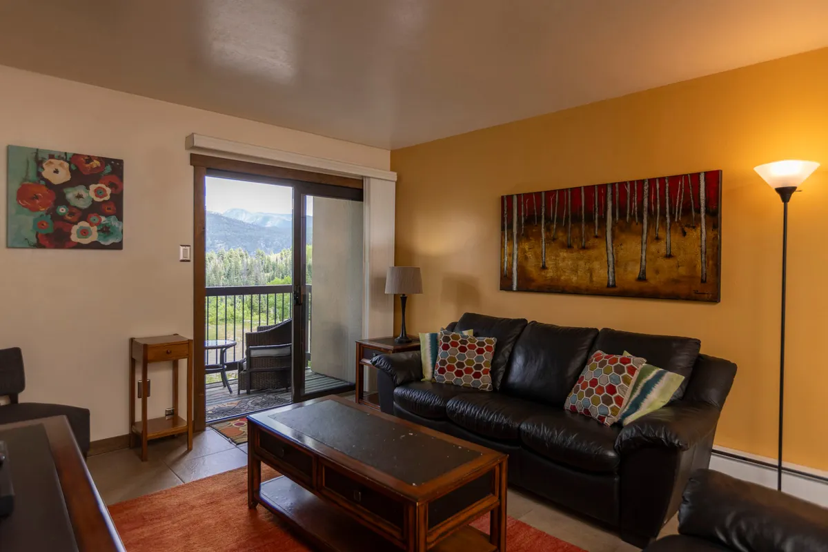 A cozy living room featuring a plush sofa with colorful cushions, artwork on the wall, and a window offering a scenic view. The ambiance is warm and inviting, ideal for relaxing and socializing after a day on the slopes.