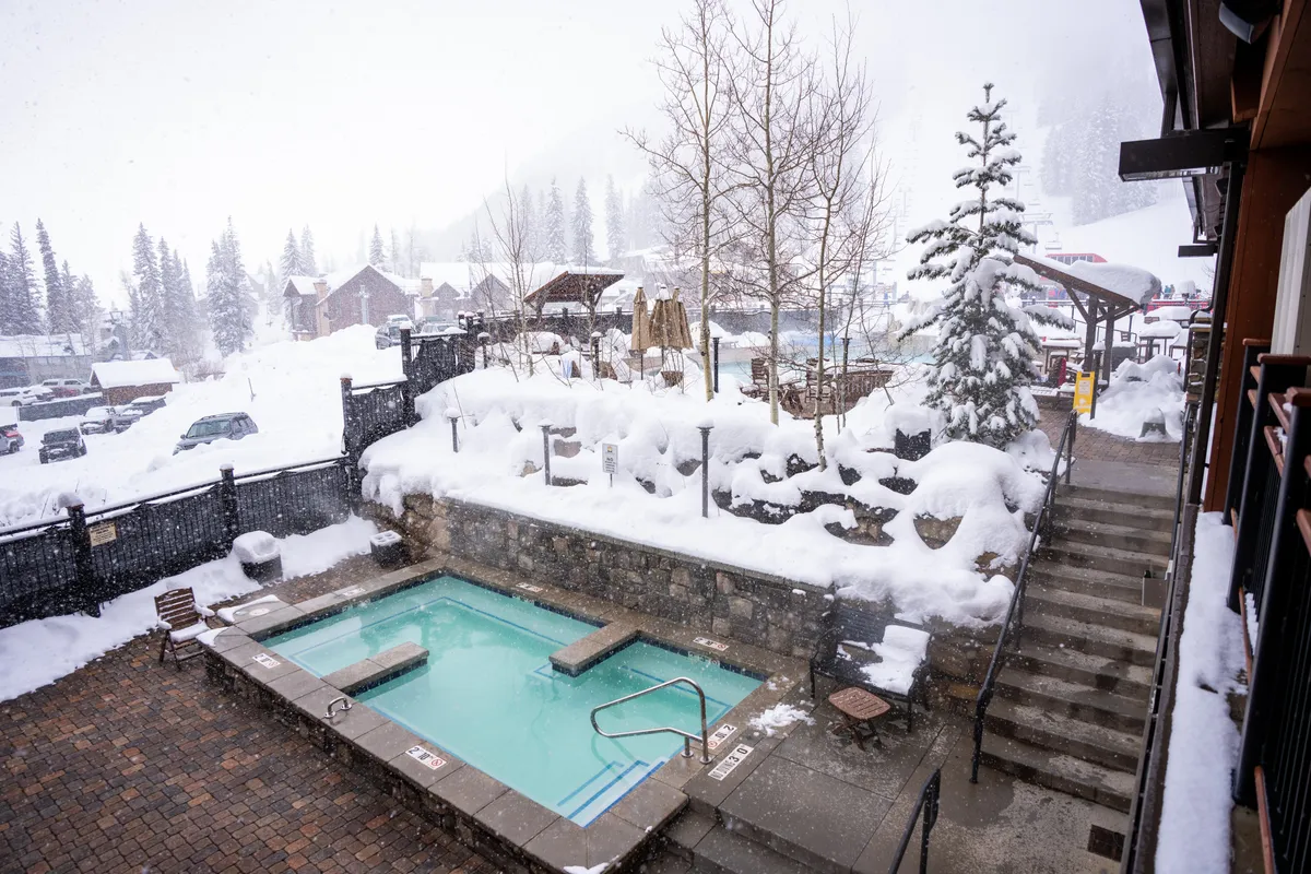 An outdoor hot tub nestled among snow-covered pines, with steam rising from the warm water. The cozy stone surroundings and steps leading up invite guests to relax and soak while enjoying a serene winter landscape.