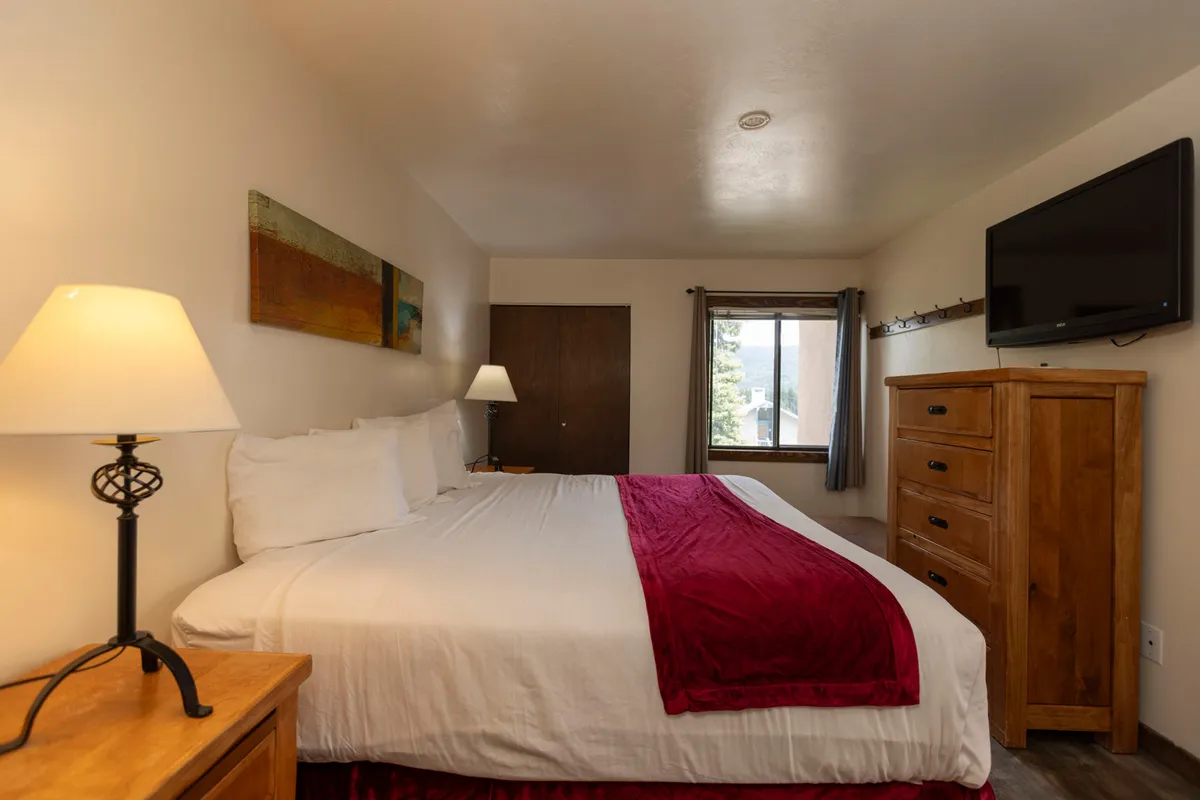 A cozy and spacious master bedroom featuring a king-size bed with crisp white linens, a wooden dresser, a reading lamp, abstract artwork above the headboard, and a large window letting in natural light. The overall mood is inviting, ideal for restful sleep and private relaxation.