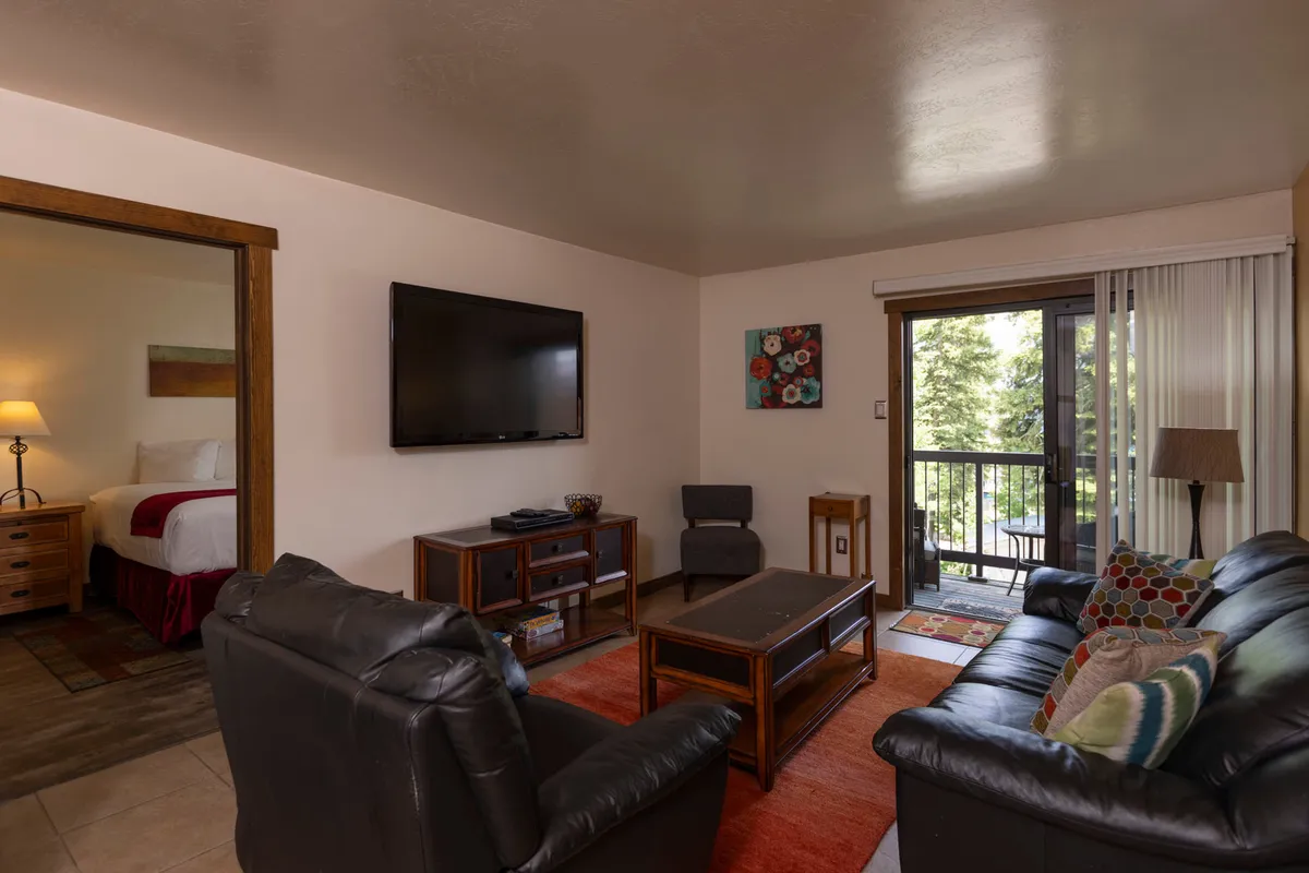 The image showcases a cozy living room with a comfortable sofa set arranged around a coffee table, a large television for entertainment, and a balcony door that floods the space with natural light. Inviting artwork adds warmth, and the setup is perfect for relaxing, socializing, or enjoying scenic views from the comfort of the indoors.