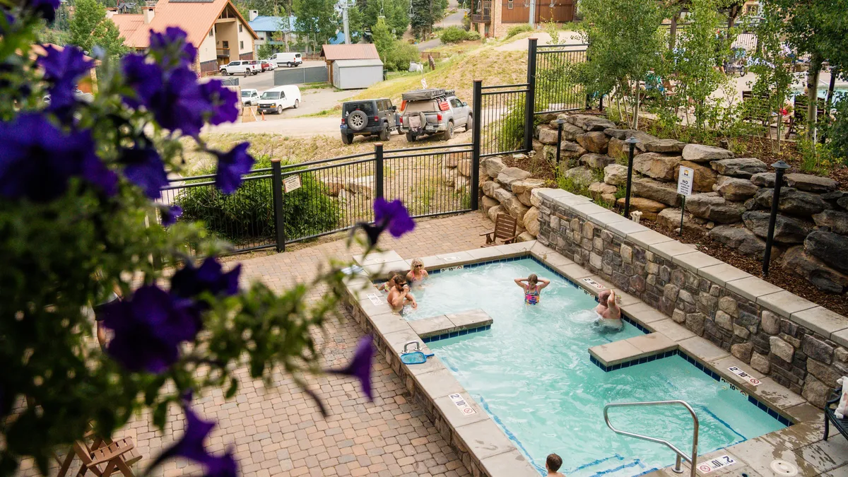A vibrant outdoor swimming pool surrounded by lush greenery and colorful flowers, with people swimming, relaxing, and socializing around the poolside. The setting feels casual, inviting, and perfect for enjoying sunny days just steps from the lodge.