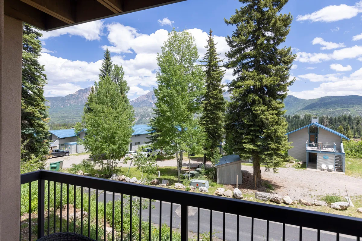 A panoramic view features green trees, a clear blue sky, and nearby resort buildings nestled in a mountain setting. The image blends natural beauty with the context of accessible amenities, suggesting proximity to shared spaces like the pool or hot tub.