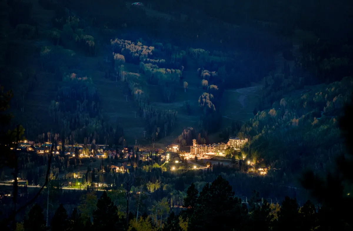 A peaceful evening scene highlights softly illuminated buildings surrounded by a tree line, with distant hills under a dusky sky. The mood is inviting and restful, suggesting family gatherings or winding down after an adventurous day.